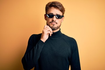 Obraz premium Young man wearing thug life fanny sunglasses standing over isolated yellow background with hand on chin thinking about question, pensive expression. Smiling with thoughtful face. Doubt concept.