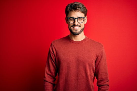 Young handsome man with beard wearing glasses and sweater standing over red background with a happy and cool smile on face. Lucky person.