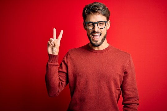 Young handsome man with beard wearing glasses and sweater standing over red background smiling with happy face winking at the camera doing victory sign with fingers. Number two.