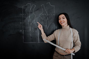 Young Asian woman seamstress points a finger at the drawing on the board, teaches sewing. Drawing of a pattern on a blackboard in a design studio for sewing and tailoring, special education concept