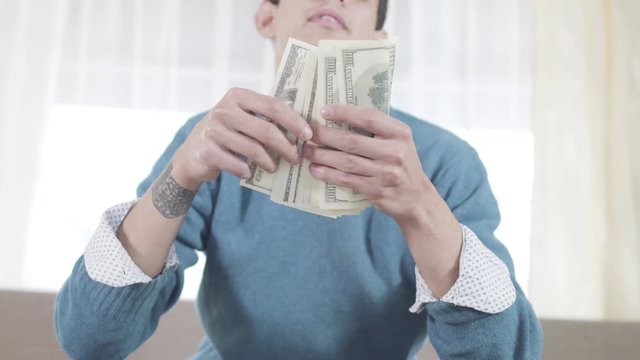 Satisfied Caucasian young man throwing up and scattering dollars. Smiling guy in eyeglasses sitting on couch and smiling. Richness, leisure, lifestyle. Camera moving up.