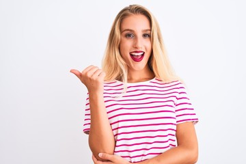 Young beautiful woman wearing pink striped t-shirt standing over isolated white background smiling with happy face looking and pointing to the side with thumb up.