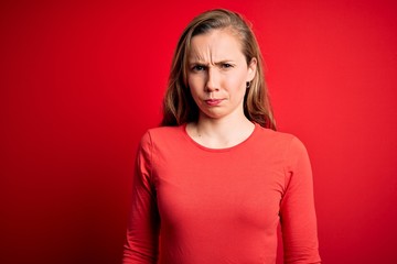 Young beautiful blonde woman wearing casual t-shirt standing over isolated red background skeptic...