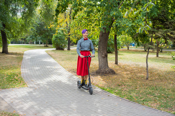 A young woman with purple hair rides an electric scooter in a park. Stylish girl in a plaid shirt, a long red skirt and a bow tie is riding around the city on a modern device. Hipster.