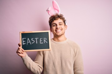 Young handsome blonde man wearing easter rabbit ears and holding blackboard with holiday word with a happy face standing and smiling with a confident smile showing teeth