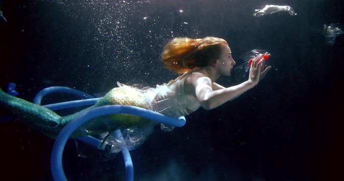 a fairy-tale mermaid girl is in a dark Studio under water. It floats among bubbles and debris. Floating long corrugated pipes, cellophane bags, plastic cups. Polluted ecology.