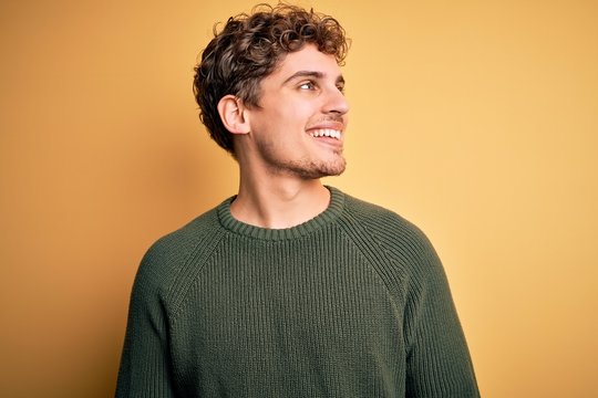 Young blond handsome man with curly hair wearing green sweater over yellow background looking away to side with smile on face, natural expression. Laughing confident.