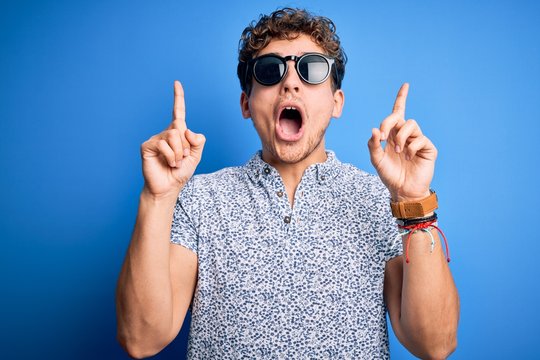 Young blond handsome man with curly hair on vacation wearing striped polo and sunglasses amazed and surprised looking up and pointing with fingers and raised arms.