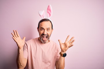 Naklejka premium Middle age hoary man wearing bunny ears standing over isolated pink background celebrating crazy and amazed for success with arms raised and open eyes screaming excited. Winner concept