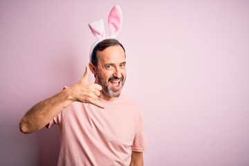 Obraz premium Middle age hoary man wearing bunny ears standing over isolated pink background smiling doing phone gesture with hand and fingers like talking on the telephone. Communicating concepts.