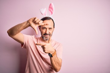 Middle age hoary man wearing bunny ears standing over isolated pink background smiling making frame with hands and fingers with happy face. Creativity and photography concept.