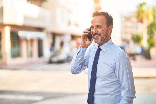 Middle age handsome businessman standing on the street talking on the smartphone smiling