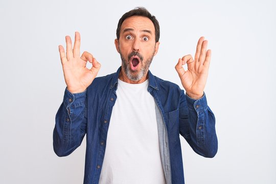 Middle age handsome man wearing blue denim shirt standing over isolated white background looking surprised and shocked doing ok approval symbol with fingers. Crazy expression