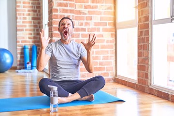Middle age handsome sportman sitting on mat doing stretching yoga exercise at gym crazy and mad...