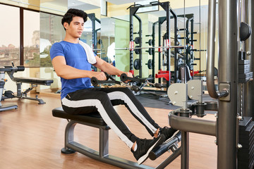 Horizontal full shot of handsome young Asian man doing exercise using fitness machine in spa resort hotel gym