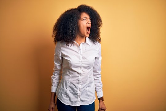 Young beautiful african american elegant woman with afro hair standing over yellow background angry and mad screaming frustrated and furious, shouting with anger. Rage and aggressive concept.