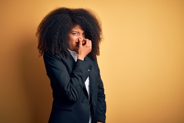 Young beautiful african american business woman with afro hair wearing elegant jacket smelling something stinky and disgusting, intolerable smell, holding breath with fingers on nose. Bad smell