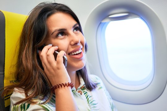 Young traveller woman sitting inside plane at the airport with sky view from the window calling using smartphone