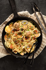 Pasta Spaghetti alle Vongole Seafood pasta with Clams, lemon and tomatoes on linen cloth in black frying pan with tongs on black background copy space