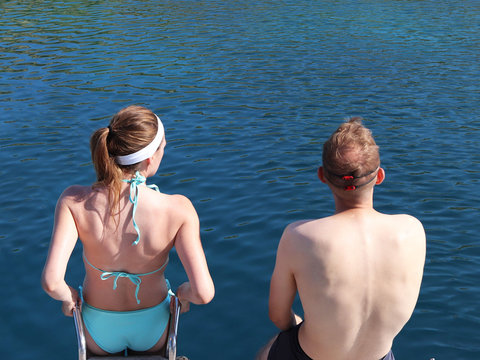 A young couple a guy and a girl in a blue swimsuit are standing on the yacht ramp and are going to take a dip in the sea. Exclusive holiday at the sea. Relax in a paradise bay. Dinghi moored