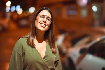 Fototapeta premium Young beautiful girl smiling happy and confident walking at the town street, standing with a smile on face around night lights bokeh
