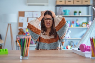 Young beautiful teacher woman wearing sweater and glasses sitting on desk at kindergarten suffering...