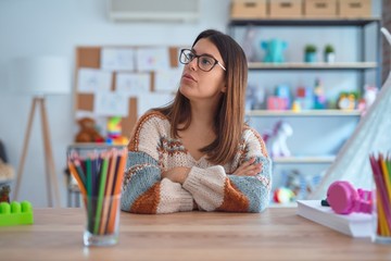 Young beautiful teacher woman wearing sweater and glasses sitting on desk at kindergarten looking...