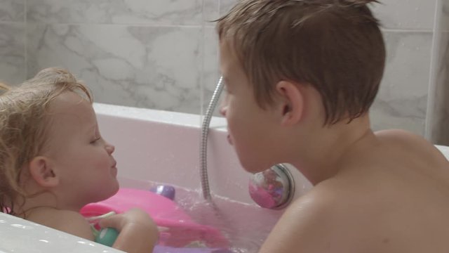 A fair haired baby girl and her elder brother are sitting in bath face to face. The boy is playfully spitting water in girls mouth. She is laughing and trying to spit it back at him