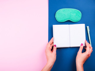 Female hands with red manicure holding open sleep journal with white pages and turquoise sleeping mask over trendy pink and classic blue background. Sleep routine, optimization concept.