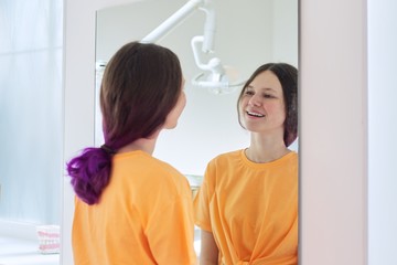 Beautiful teenager girl looking at white healthy teeth in mirror in dentist office