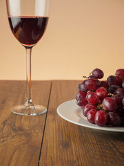 Glass of red wine and plate with red grapes on a wooden table, Warm light background. Grapes are in focus.