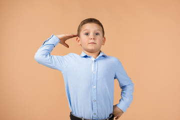 Cute little child boy in blue shirt showing a saluting gesture on beige background. Human emotions and facial expression