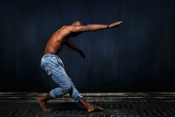 A black athletic man in jeans with a naked torso is dancing against a dark background.