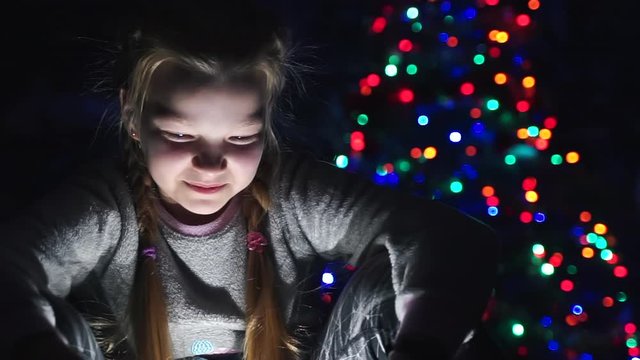 laughing girl watching xmas cartoon at dark night. Christmas tree bokeh magic lights background. child watching funny video movie on tablet looking at screen. Happy 2021 New year. Winter vacation home