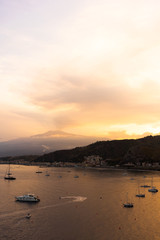 view of Mount Etna volcano with a town, port and boats below it during sunset (Sicily, Italy) 