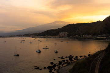 view of Mount Etna volcano with a town, port and boats below it during sunset (Sicily, Italy) 