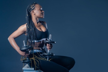 Fototapeta na wymiar African-American model with dumbbells on a dark background. Girl with braids involved in sports. Black athlete in a fitness club. Sale of goods for sports. Sports equipment. Healthy lifestyle.