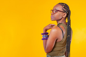 African student wearing glasses on a yellow background. Ethnic flavor.Dark-skinned girl with braids. Original jewelry. Advertising hairdressing services. Place for inscription. Purple fringed bracelet