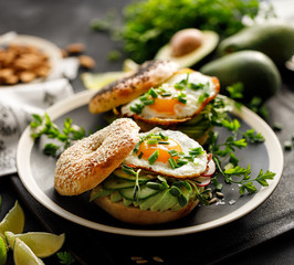 Bagel sandwich with avocado, fried egg, fresh cucumber, radish and sunflower sprouts on a black plate, close-up view. Vegetarian food