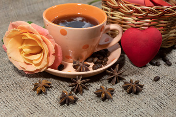 On the table is a cup of coffee. On a saucer lies a rose. Behind is a basket and a red heart. Near scattered coffee beans and star anise.