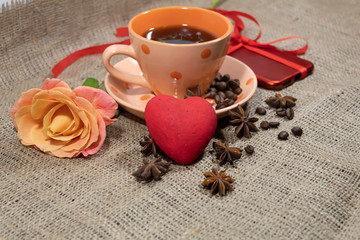 On the table is a cup of coffee. Ahead lies a rose, a red heart and a smartphone is visible. On a saucer and on a table are coffee beans and star anise.
