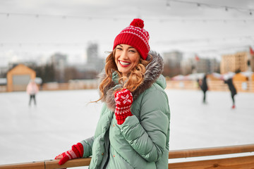 Beautiful lovely middle-aged girl with curly hair warm winter jackets stands ice rink background Town Square.