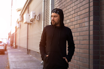 Handsome young unshaven man black hoodie and bandana stands backyard near brick walls summer day.