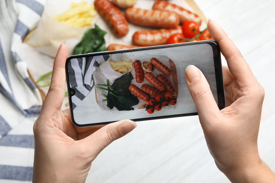 Blogger taking photo of grilled sausages with garnish at white table, closeup