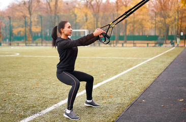 Girl athlete functional training on sportground. Mixed race young adult woman do workout with suspension system. Healthy lifestyle. Stretching outdoors playground.