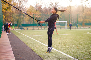 Girl athlete functional training on sportground. Mixed race young adult woman do workout with suspension system. Healthy lifestyle. Stretching outdoors playground.
