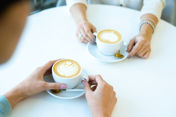 Hands of two friendly girls having fresh tasty cappuccino during communication