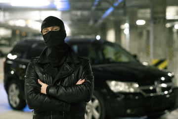 Male criminal in black jacket and balaclava standing on parking area