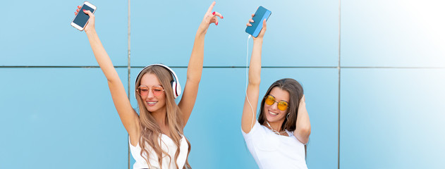 Fashion portrait of two pretty smiling hipsters woman in sunglasses holding smartphone and dancing...