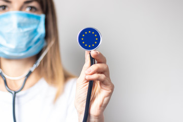 Young woman in a medical mask holds a stethoscope with the flag of European Union on a light background. Concept of medicine, virus, epidemic, vaccination © Alex